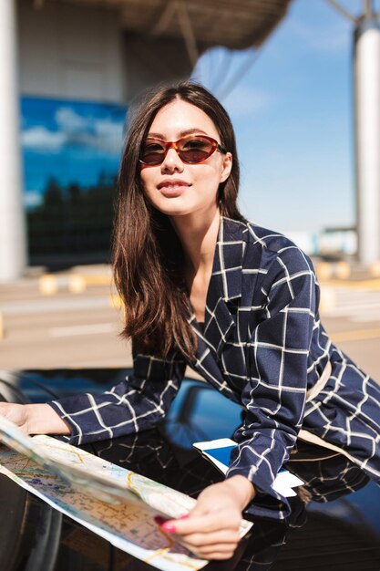 Красивая девушка в солнечных очках с картой в руках, опираясь на машину, мечтательно глядя в камеру на фоне аэропорта