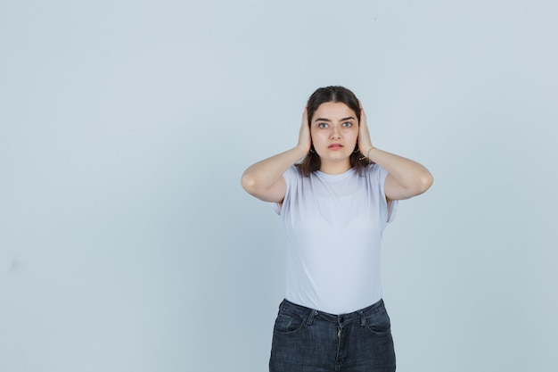 Bella ragazza in t-shirt, jeans che tengono le mani sulle orecchie e guardando frustrato, vista frontale.