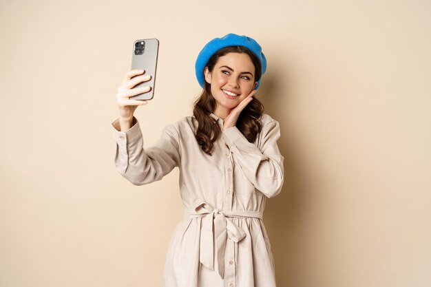 Beautiful girl taking selfie on smartphone in coat and cute hat, smiling for photo and looking happy, standing over beige background