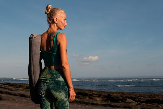Красивая девушка гуляет по пляжу с ковриком для йоги