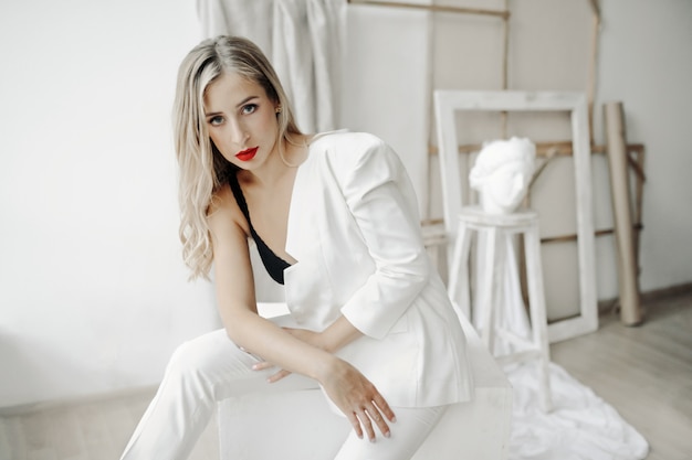 Beautiful girl wearing in a bra and a white suit on her one shoulder sits on a white cube