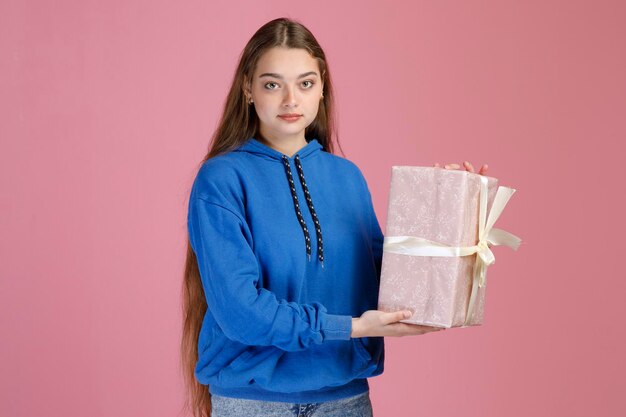 Красивая девушка в повседневной одежде держит подарок, завернутый в глянцевую бумагу, серьезно смотрит в камеру