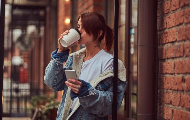 데님 코트를 입은 아름다운 소녀가 카페 밖에서 스마트폰을 들고 테이크아웃 커피를 마십니다.