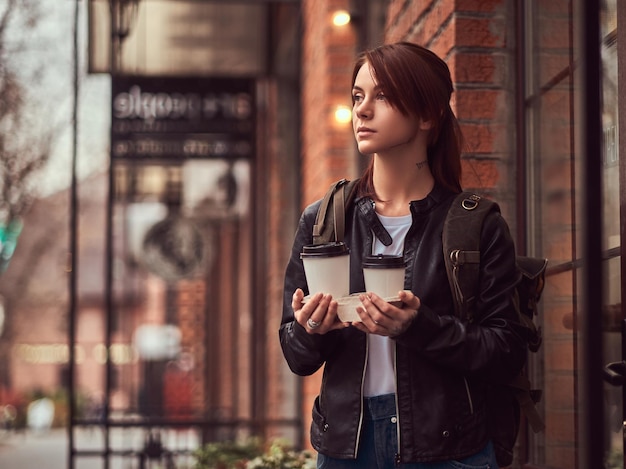 A beautiful girl wearing a leather jacket with a rucksack holding cups with takeaway coffee outside near the cafe.