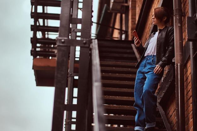 A beautiful girl wearing trendy clothes standing on stairs outside the cafe and holding a smartphone.