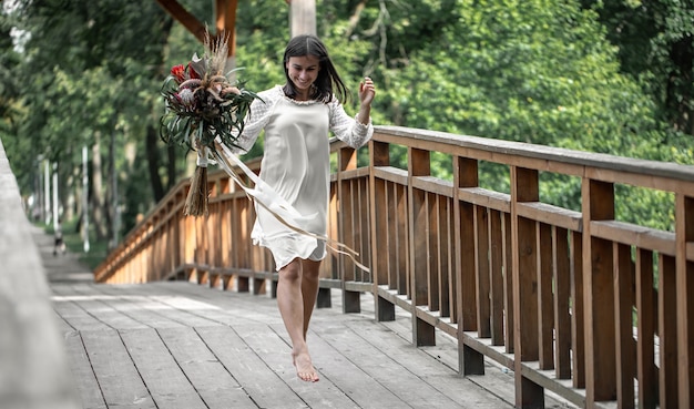 Красивая девушка в белом платье с букетом экзотических цветов на деревянном мосту.