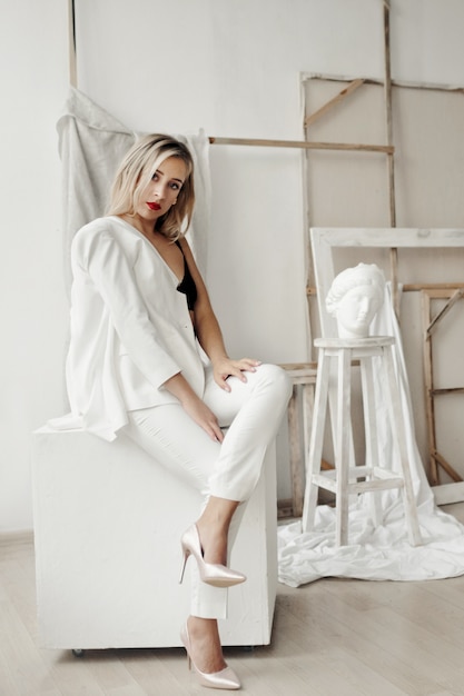 Beautiful girl in a white suit sits on a white cube in a gallery