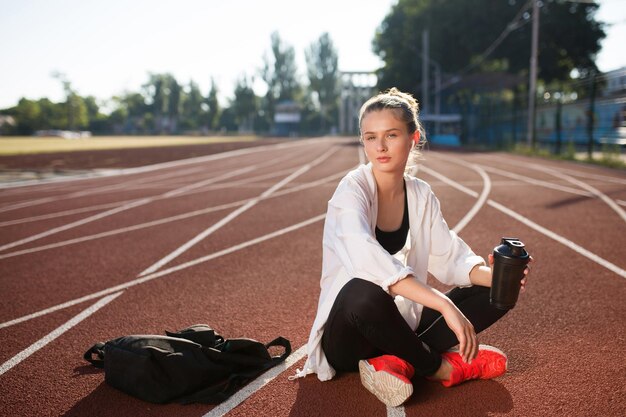 Beautiful girl in wireless earphones holding sport bottle in hand dreamily looking in camera while spending time on running track of stadium