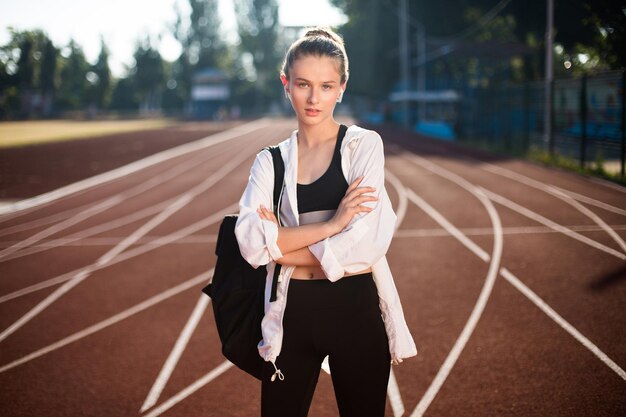 Beautiful girl in wireless earphones with backpack on shoulder dreamily looking in camera on treadmill of city stadium