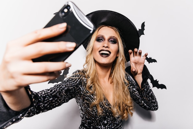 Beautiful girl in witch attire making selfie at carnival. Long-haired blonde lady chilling in halloween.