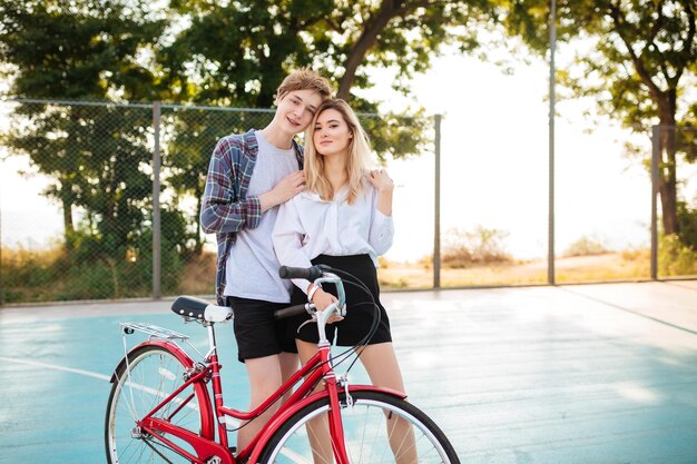 Beautiful girl with blond hair happily looking in camera while standing with happy boy and holding his hand in park Portrait of cute young couple embracing one another with classic red bicycle near