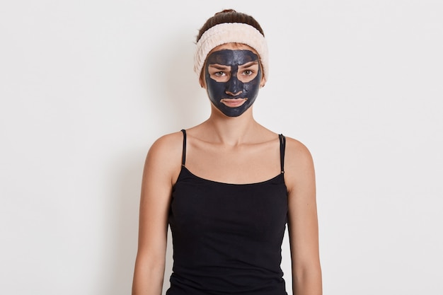 Красивая девушка с глиняной маской на лице стоя с расстроенным выражением лица с грустью, носить черную футболку и ленту для волос.