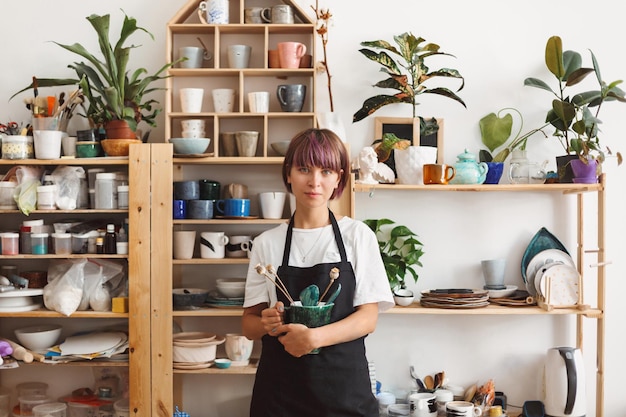Бесплатное фото Красивая девушка с разноцветными волосами в черном фартуке и белой футболке держит миску ручной работы с растением в руках, мечтательно глядя в камеру в гончарной мастерской
