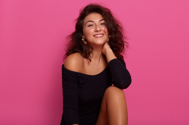 Beautiful girl with dark wavy hair, sitting isolated over pink studio, keeps hand on neck, posing with bare shoulders