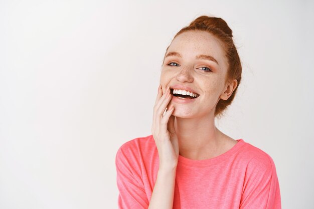 Beautiful girl with ginger hair combed in bun touching clean and healthy skin, smiling happy at front, standing in pink t-shirt over white wall