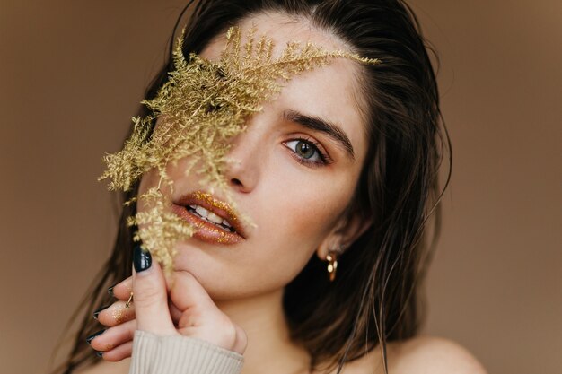 Beautiful girl with golden earrings posing with plant. Indoor photo of blissful brunette woman with flower.