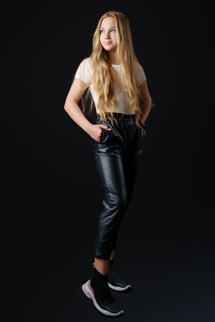 A beautiful girl with long blonde hair in a white t shirt and black leather pants poses for the camera on a black background