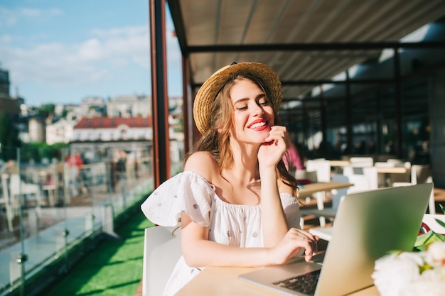 Beautiful girl with long hair in hat sits at table on the terrace in cafe . She wears a white dress with bare shoulders and red lipstick . She looks happy typing on laptop.