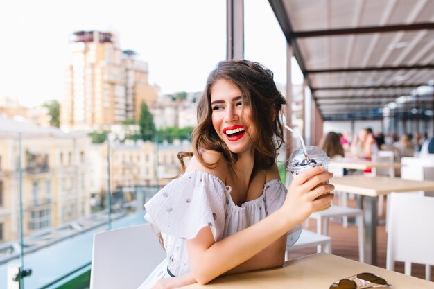 Beautiful girl with long hair is sitting at  table on the terrace in cafe . She wears a white dress with naked shoulders and red lipstick . She holds cup to go and smiles to the side.