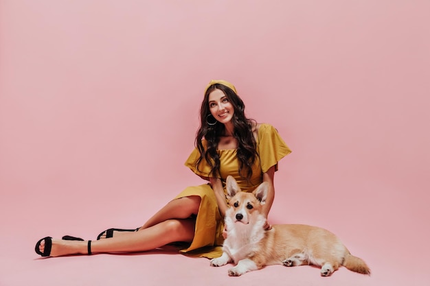 Free photo beautiful girl with long hair in modern yellow dress and dark shoes smiling and posing with corgi on pink isolated background