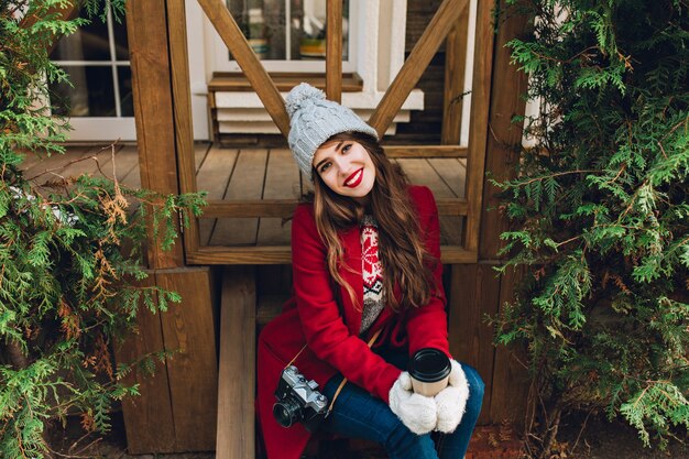 Beautiful girl with long hair in red coat sitting on wooden stairs between green branches outdoor. She holds coffee in white gloves and smiling . View from above.