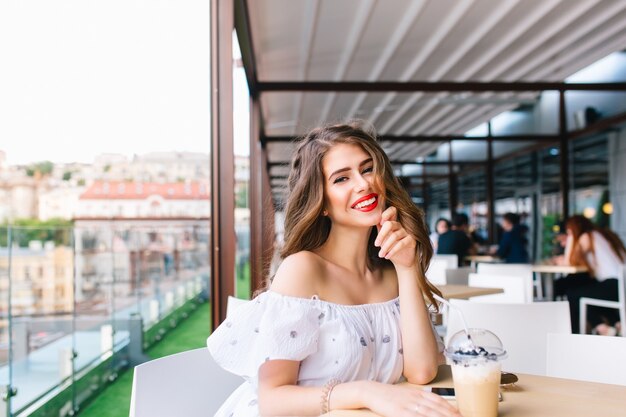 Красивая девушка с длинными волосами сидит за столом на террасе в кафе. Она носит белое платье с открытыми плечами и красной помадой. Она улыбается в камеру.