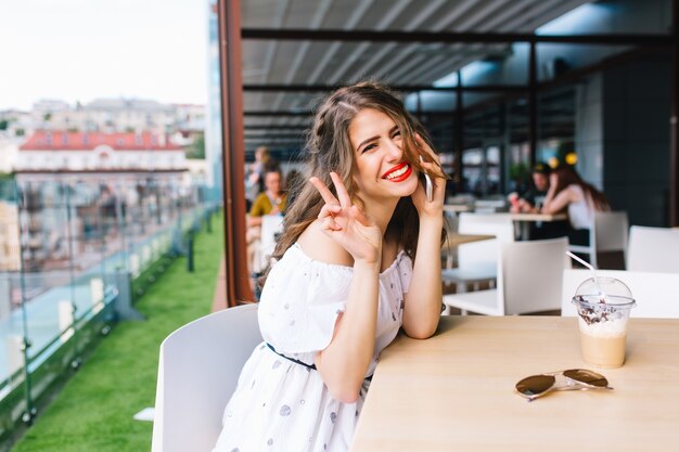 Beautiful girl with long hair sits at  table on the terrace in cafe . She wears a white dress with bare shoulders and red lipstick . She is speaking on phone and smiling to the camera.