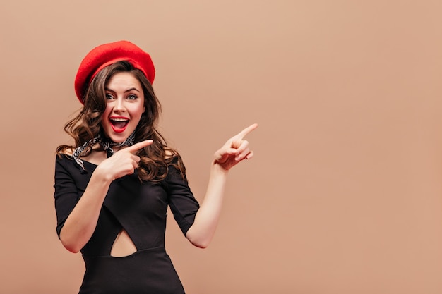 Красивая девушка с волнистыми волосами улыбается и указывает пальцами вправо. портрет женщины в красной шляпе и черном элегантном платье на бежевом фоне.