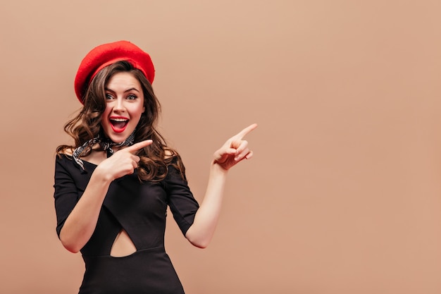 Beautiful girl with wavy hair smiles and points with her fingers to right. Portrait of woman in red hat and black elegant dress on beige background.