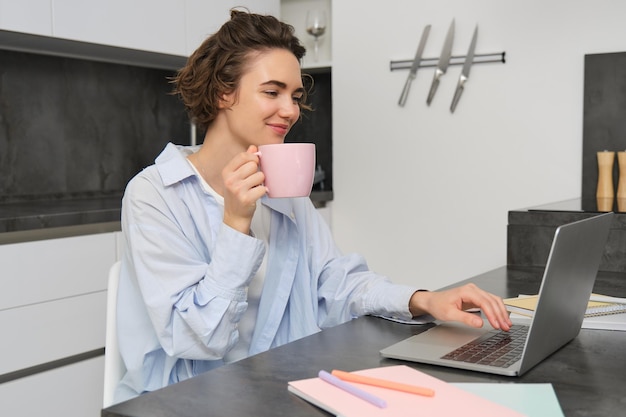 Foto gratuita bella ragazza che lavora da casa bevendo caffè e guardando il portatile, seduta una giovane donna d'affari