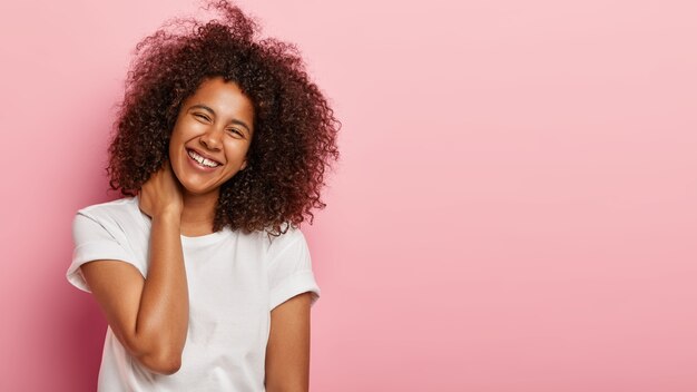 Beautiful girlfriend with funny look has nice talk, touches neck gently, laughs out happily at hilarious joke, being in excellent mood dressed in white t shirt stands against pink wall with copy space