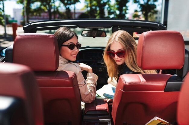 Beautiful girls in sunglasses happily looking in camera spending time together while driving cabriolet car on city streets outdoor