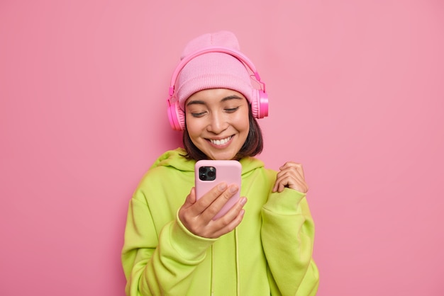 Free photo beautiful glad asian female teenager looks happily at smartphone listens music via headphones enjoys favorite playlist wears hat and green sweatshirt isolated over pink wall