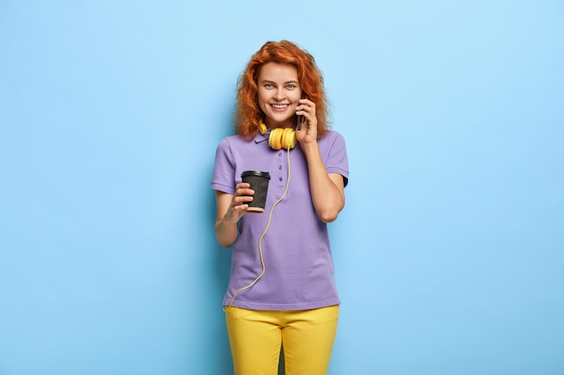 Free photo beautiful glad female has funny conversation, uses modern cell phone, drinks takeaway coffee