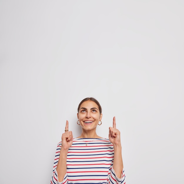 Beautiful glad young woman with cheerful expression happy smile on face points index fingers above found nice product shows promo dressed in casual striped jumper isolated over white background