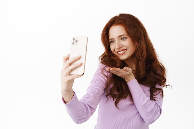 Beautiful glamour girl with red hair sending air kiss at smartphone camera, video calling lover, smiling flirty, taking cute selfie on mobile phone, standing against white background