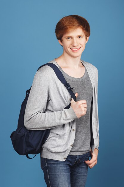 Красивый красивый веселый студент с рыжими волосами в повседневной одежде улыбается, держит рюкзак и рука в кармане, с расслабленным и счастливым выражением.