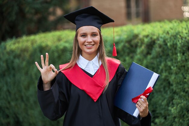 Beautiful graduate girl in graduation robe shows ok sign and smiling at camera.