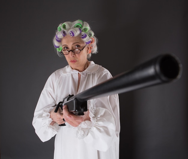 Free photo beautiful grandmother posing with weapon in studio. serious lady with rollers on looking at camera and holding rifle.