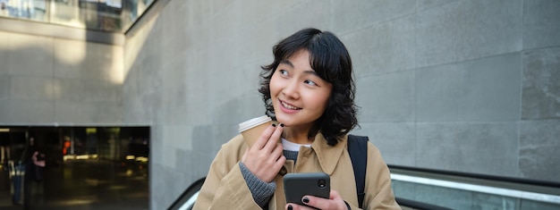 無料写真 美しい幸せなアジアの女の子は、エスカレーターに立ちながら携帯電話を使用してコーヒーを飲みます