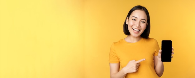 Beautiful happy asian girl showing mobile phone screen application on smartphone gadget standing ove