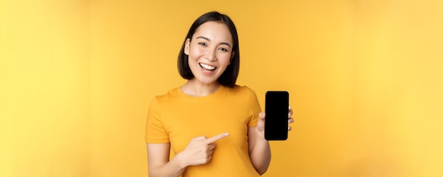 Beautiful happy asian girl showing mobile phone screen application on smartphone gadget standing ove