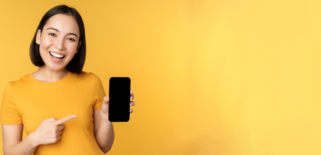 Beautiful happy asian girl showing mobile phone screen application on smartphone gadget standing over yellow background