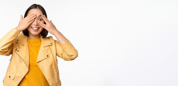 Beautiful happy asian woman shuts eyes blindfolded waiting for surprise and smiling peeking through fingers anticipating gifts on holiday standing over white background