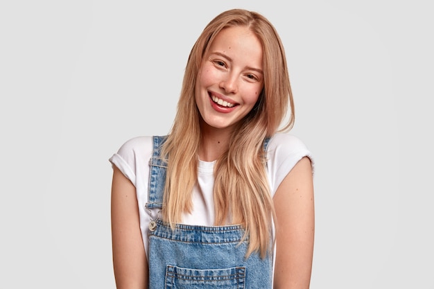 Beautiful happy Caucasian female with charming smile, dressed in fashionable overalls, has long light hair, expresses positive emotions as sees something pleasant in front, stands against white wall