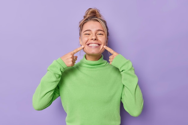 Free photo beautiful happy fair haired european woman points index fingers at mouth forces cheerful smile shows perfect white teeth wears green jumper being in good mood isolated over purple background.