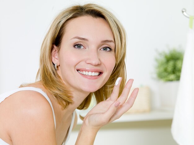 Beautiful happy girl applying moisturizer  cream on face standing in the bathroom