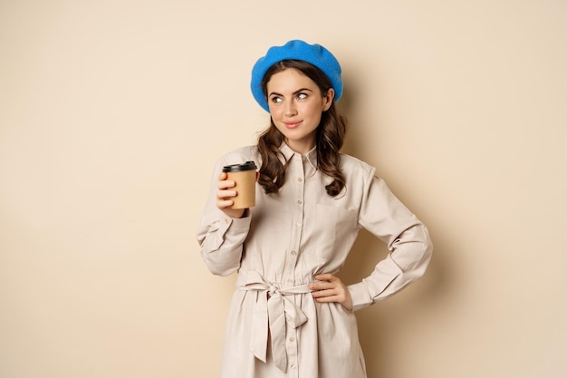 Красивая счастливая девушка пьет кофе на вынос из кафе и улыбается, позирует с чашкой напитка, бежевый фон