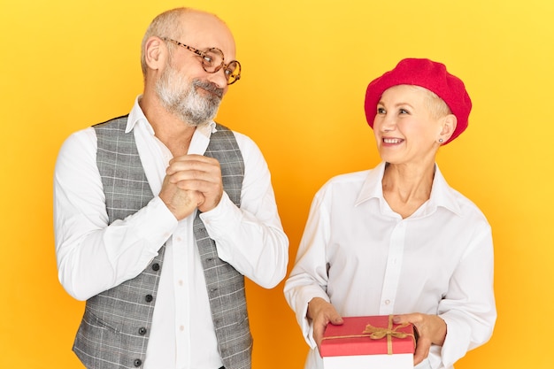 Foto gratuita bella donna matura felice in berretto rosso che riceve il regalo di compleanno dal marito che si congratula con lei con tutto il cuore. uomo colpevole e triste che ripara la sua colpa, conquistando la moglie con un regalo