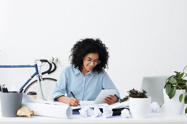 Free photo beautiful happy mixed race female architect smiling, writing down calculations from digital tablet